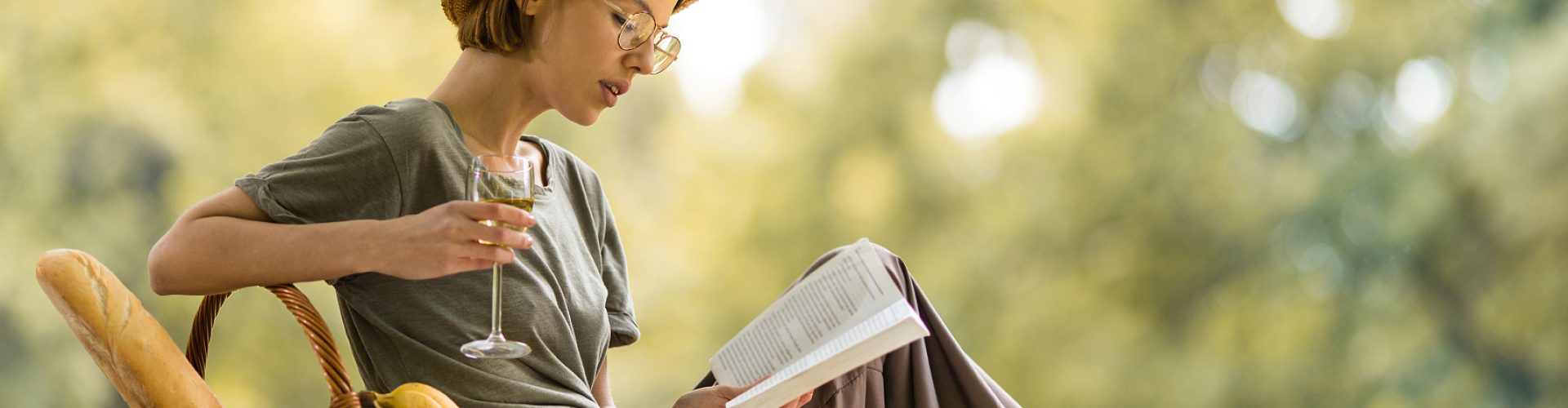 Relaxed woman enjoying in nature at springtime and reading a book while drinking white wine.