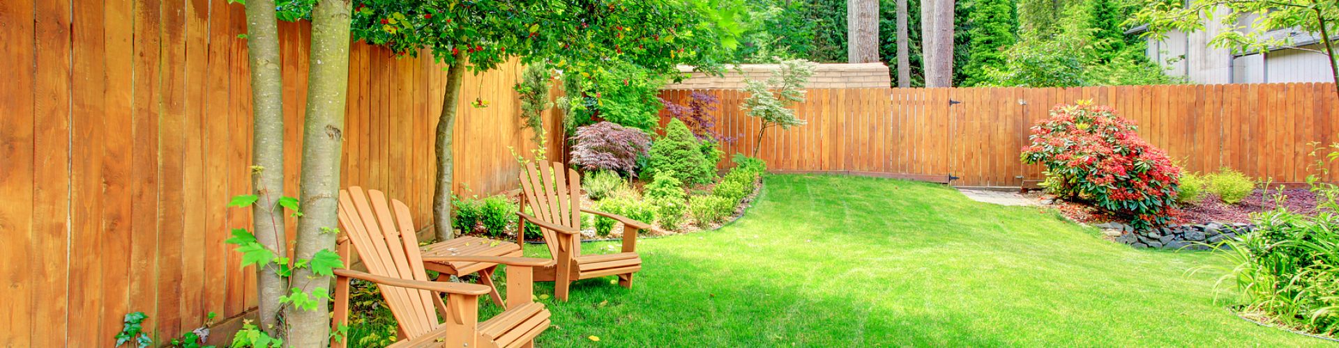 Fenced backyard with green lawn, flower beds and romantic sitting area with wooden chairs and table