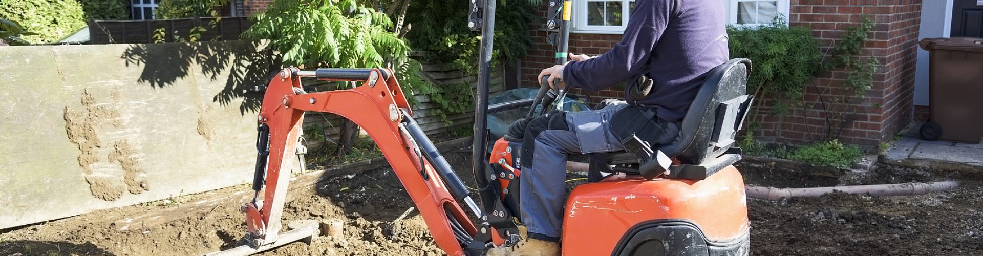 A white Caucasian male is operating a small orange digger, turning up soil and earth as ground works for a new driveway in front of a house. He is wearing casual clothing, a fleece and a wooly hat. The digger arm is extended into the earth he is digging.